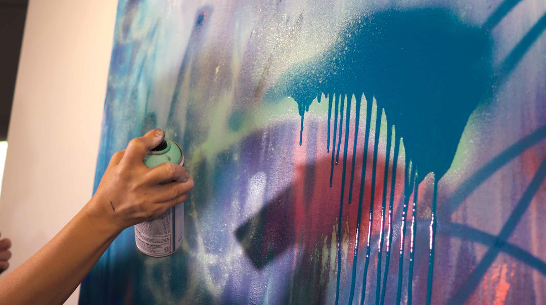 Elli's hand holds a can of blue spray paint, which makes drips on a messy canvas