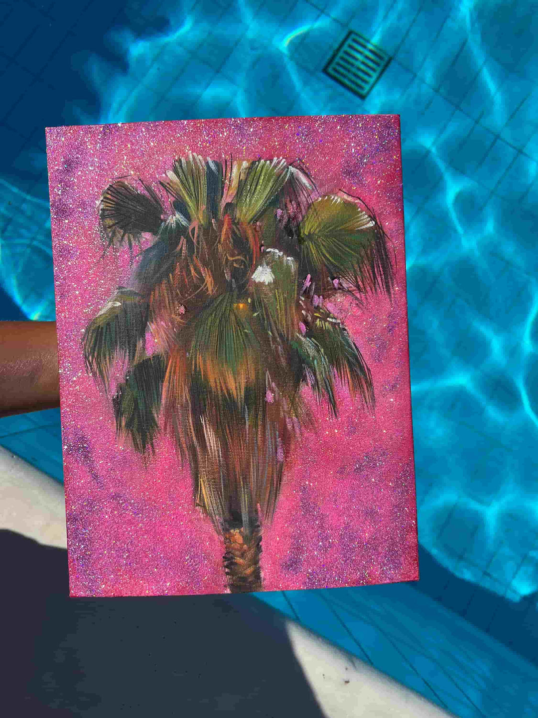An overhead shot of a painting of a palm tree with a pink background as it is held above a sparkling blue swimming pool