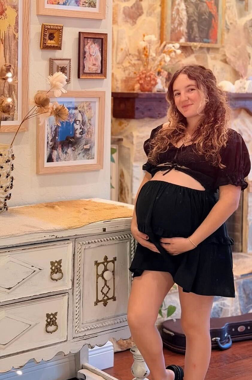 Pregnant Dimitra poses near a wall of artwork in her home