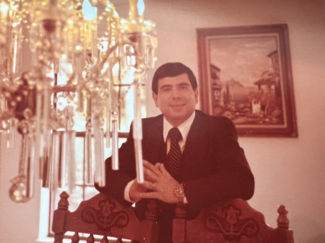 Elli Milan's greek father, George, dressed in a dapper suit, smiling in a dining room with a crystal chandelier