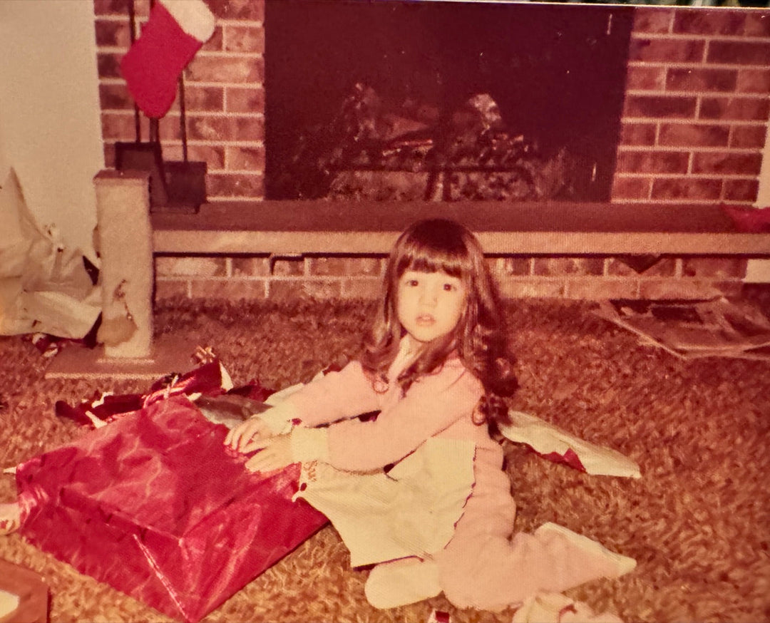 Elli Milan as a little girl opening a red-wrapped present on Christmas morning by a brick fireplace